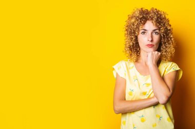Düşünceli kıvırcık kadın sarı arka planda çenesinin altında bir yumruk tutuyor.