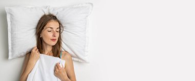 Beyaz bir yatakta uyuyan genç bir kadın. Üst manzara, düz yatış. Pankart.