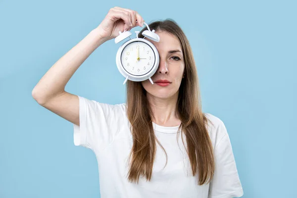 Sleepy young woman covering her eye with alarm clock on blue background.