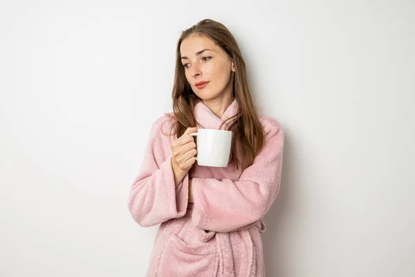 Young woman in a bathrobe drinks coffee on a white background.