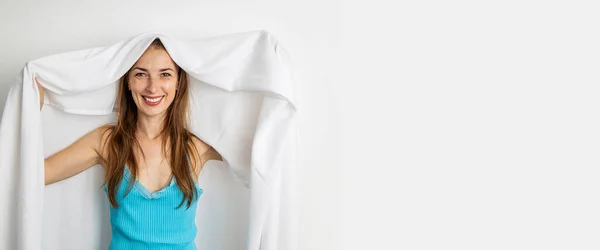 Smiling young woman covering herself with white sheet on white background.