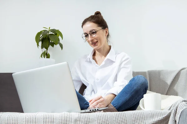 Smiling young woman with laptop with coffee sitting on sofa