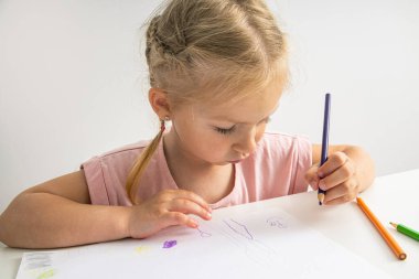 A child blonde girl draws with colored pencils sitting at the table.