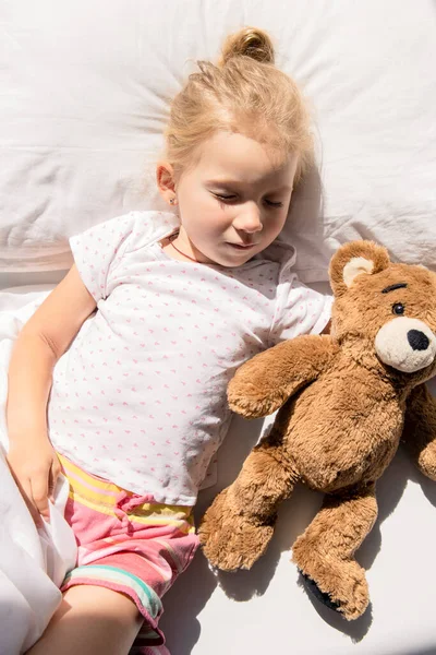 Little girl sleeps hugging a soft toy bear on a white bed. Top view, flat lay.