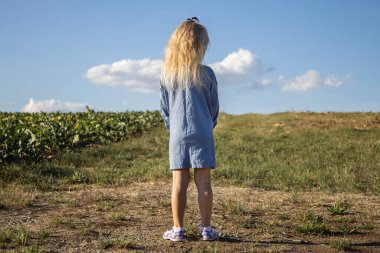 Sırtı tarlada dikilen, elbiseli sarışın bir kız..