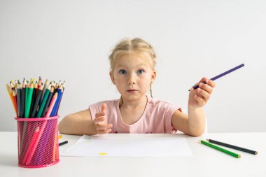 Blue-eyed blonde child girl draws with colored pencils sitting at a white table.