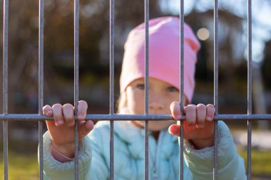 Little girl child looks through the iron fence on the street.