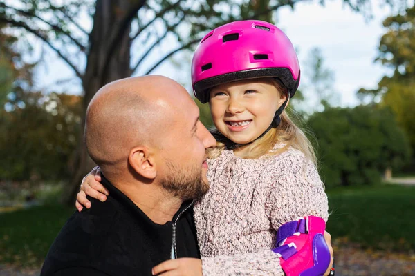 Joyful dad and little daughter in a helmet hugging in the park.