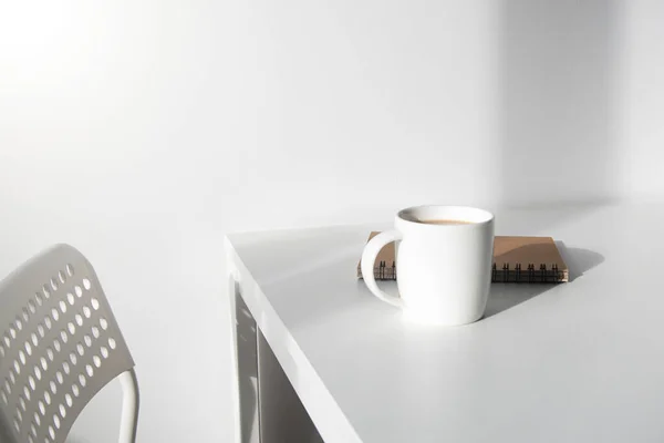 White cup of coffee, a notebook on a white table, there is a chair next to it