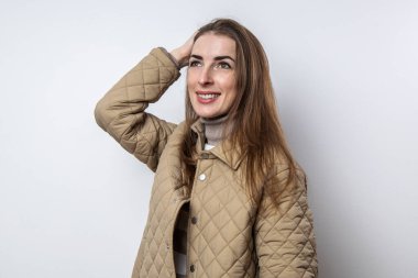 Ceketli gülümseyen kız beyaz bir duvarın yanında saçını düzeltiyor.