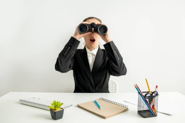 Surprised young woman looking through binoculars while sitting at desk at workplace.