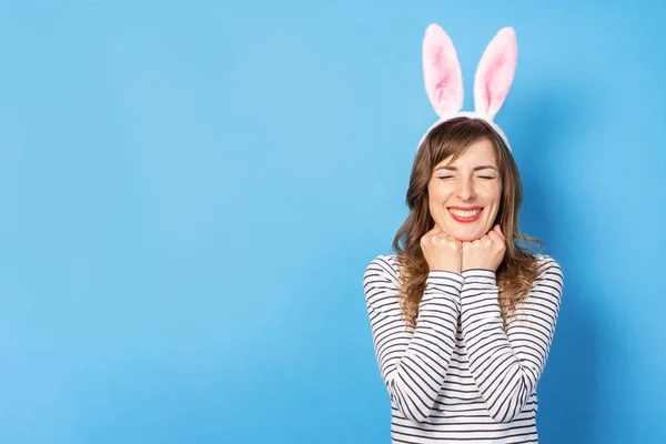 Joyful Young brunette woman with bunny ears and closed eyes holds two hands under her chin on a blue background. Easter concept, surprise, hide and seek, emotional. Face expression. Banner.