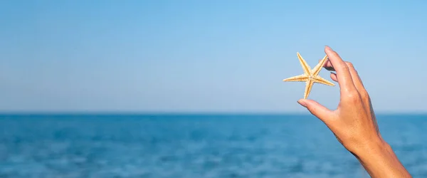 Female hand holds a starfish against the background of the sea, sky. Banner.