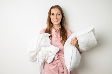 Bornozlu, elinde yastık ve beyaz bir arka planda çarşaf tutan gülümseyen genç bir kadın..