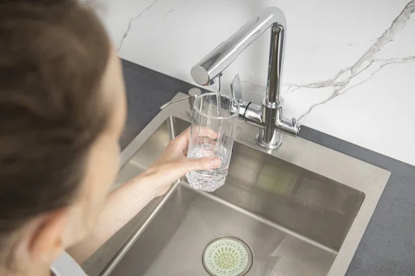 stock image Young woman holding a glass pours water from a faucet in the kitchen.