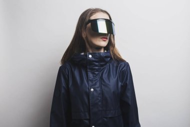 Young woman in virtual reality glasses and in a blue jacket on a light background.