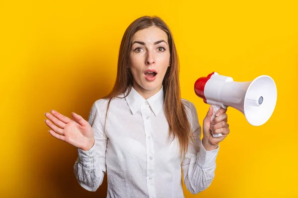 stock image Surprised young woman in a white shirt holds a megaphone on a yellow background. Hiring concept, ad.