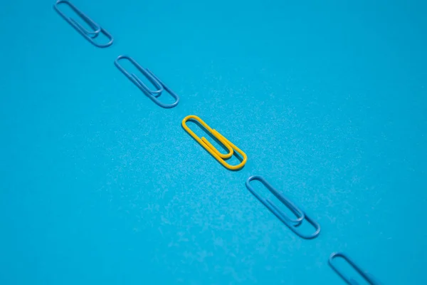 One yellow paper clip among blue paper clips on a blue background. Top view, flat lay.