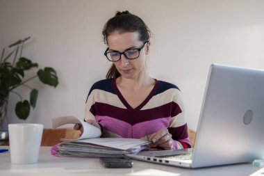 Gözlüklü genç bir kadın dizüstü bilgisayarda otururken gazetelere bakıyor..