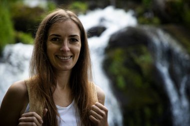 Sırt çantalı gülümseyen genç bir kadın şelalenin yanında duruyor..