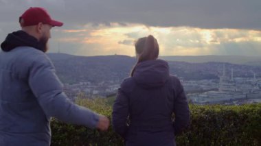 Genç bir adam şehrin panoramasında genç bir kıza sarılıyor..