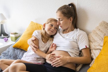 Mutlu hamile kadın kanepeye oturmuş, kızı karnını okşuyor..