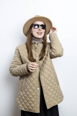 Ceketli, güneş gözlüklü, şapkalı genç bir kadın beyaz bir duvarın arkasına bakıyor.