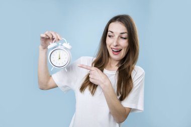 Gülümseyen genç bir kadın mavi arka plandaki çalar saati işaret ediyor..
