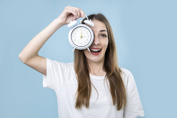 Surprised young woman covering her eye with alarm clock on blue background.