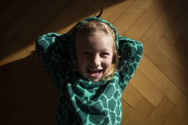 Smiling child girl in dinosaur costume lies on the floor. Top view, flat lay.