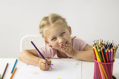 A smiling child blonde girl draws with colored pencils sitting at a white table.