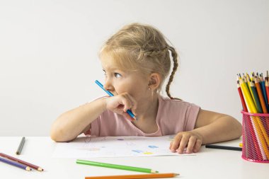 A blonde child girl draws with colored pencils sitting at a white table.