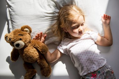 Küçük kız, güneş ışınları altında yumuşak bir oyuncak ayıyla yataktan kalkar. Üst manzara, düz uzanma.