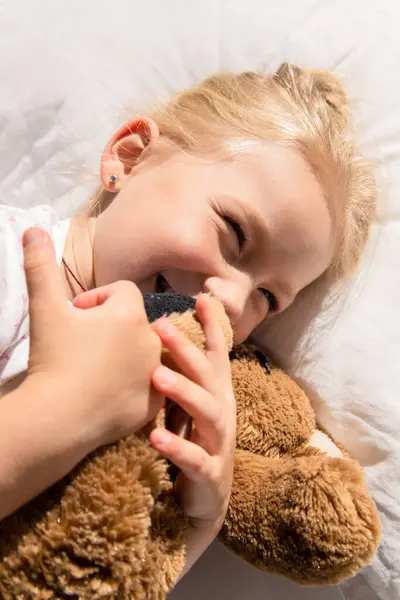 Smiling little girl hugging a soft bear lies on a white pillow. Top view, flat lay.