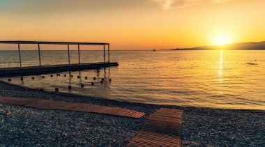 Deniz yüzeyinde ve taş plajda sarı güneşli gün batımı.