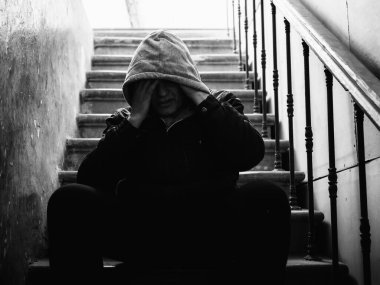 Lonely man in hood with his head down sits on stairs in dark. Stress and depression mental problems concept.
