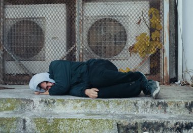 Frozen homeless man sleeps on concrete near buildings ventilation system. Problem of how homeless survive winter cold.