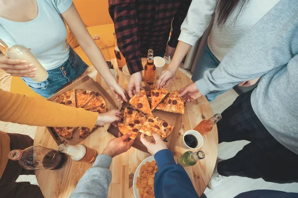 Top view of many friends hands take slices of Italian pizza at wooden table. Home party with beer bottles, cocktails and pizza from food delivery.