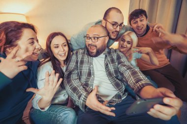 Group of young people taking selfie on smartphone at home party and have fun. Friends enjoy their company, smiling.