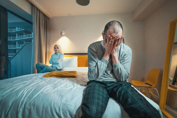 Quarrel of married couple. Man sits on edge of bed and holds his head with hands. Wife or angry girlfriend sitting cross-armed at other end of bed. Problems in relationships.