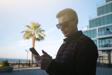 Man in plaid shirt and sunglasses uses smartphone on street against backdrop of palm trees by sea and new buildings.