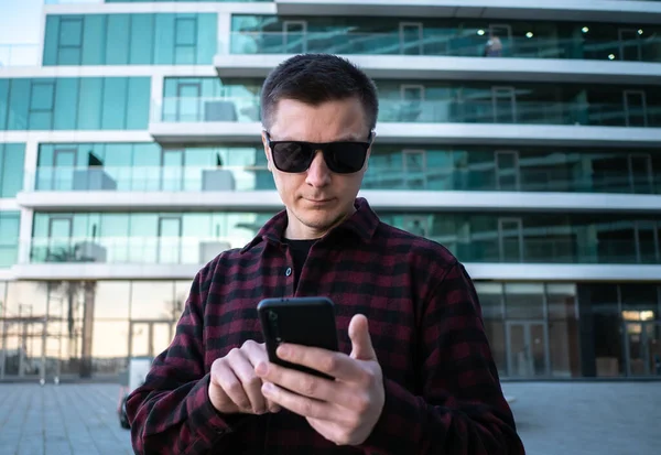 Young man in sunglasses using mobile phone in hands at modern new office building background.