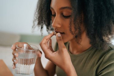 Genç Afrikalı Amerikalı kız tablet ya da ilaç kullanıyor ve elinde bir bardak su tutuyor. Sağlıklı yaşam tarzı, vitamin diyeti..