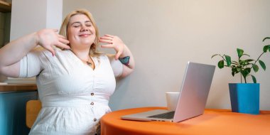 Mutlu gülümseyen, iri yarı, dizüstü bilgisayarlı bir kadın iş yerinde kahve molası veriyor..