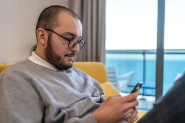 Kanepede oturan adam cep telefonuna bakıyor. Akıllı telefondan e-postaları kontrol eden ya da sosyal medyada haber okuyan adam..