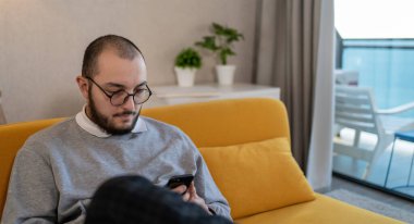 Yuvarlak gözlüklü genç adam evde akıllı telefon cihazı kullanıyor, mesaj yazıyor ya da arkadaşlarıyla sohbet ediyor..