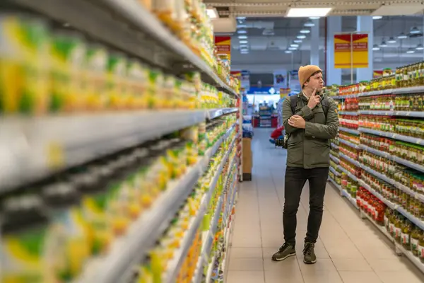 Focused young man stands contemplating his shopping and choose what to buy options in brightly lit aisle of grocery store