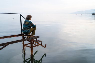 Dalgın adam, yıpranmış iskelede, sakin denizin kenarında, alacakaranlıkta, ufkun ötesine uzanan.