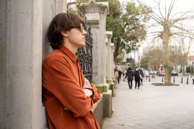 90 'ların moda tarzı ve klasik gözlüklü turuncu ceket giyen genç adam City Caddesi' ndeki sütunun yanında düşünceli duruyor. Modern geçmişe dönük ve nostaljik moda kavramı.