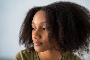 Doğal kıvırcık saçlı genç Afrikalı Amerikalı bir kadın, düşünceli görünüyor, açık bir arka planı var.. 
